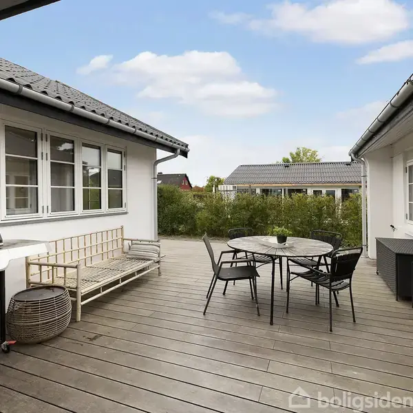 Et udendørs terrasseområde med trædæk indeholder en gasgrill til venstre, et rundt bord med stole i midten og en bænk langs huset. Omkring er der grønne buske og nabohuse.