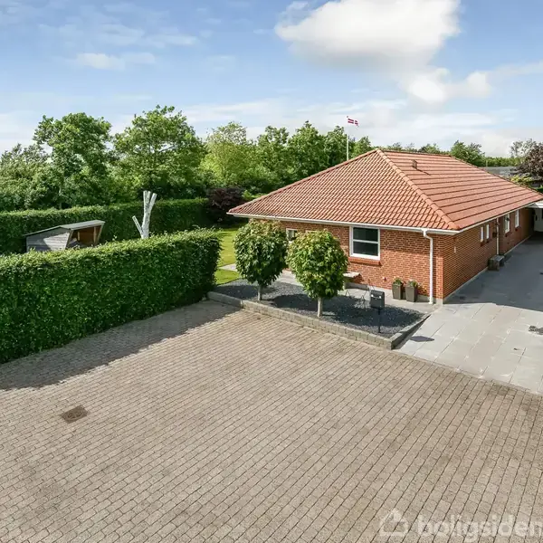 Et rødt murstenshus med et tegltag står i en have. Huset har en carport, omringet af grønne hække og flisebelagt terrasse. Himlen er blå, og der er få skyer.