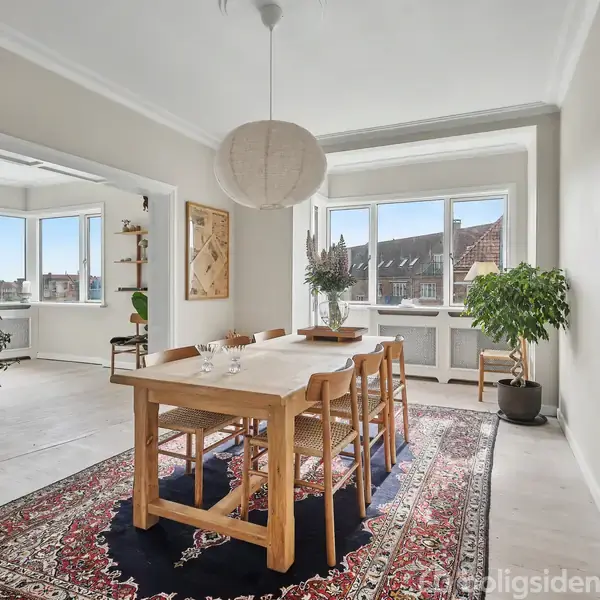Spisebord er centreret på et orientalsk tæppe, omgivet af stole. Store vinduer i baggrunden giver udsigt til hustage. Grønne planter og en stor pendellampe kompletterer den lyse spisestue.