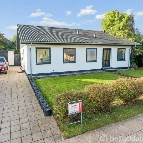 Hvid bungalow med sort tag står stille i en velholdt have. Til venstre er en indkørsel med en parkeret bil, og der er et skilt fra "home" på plænen.