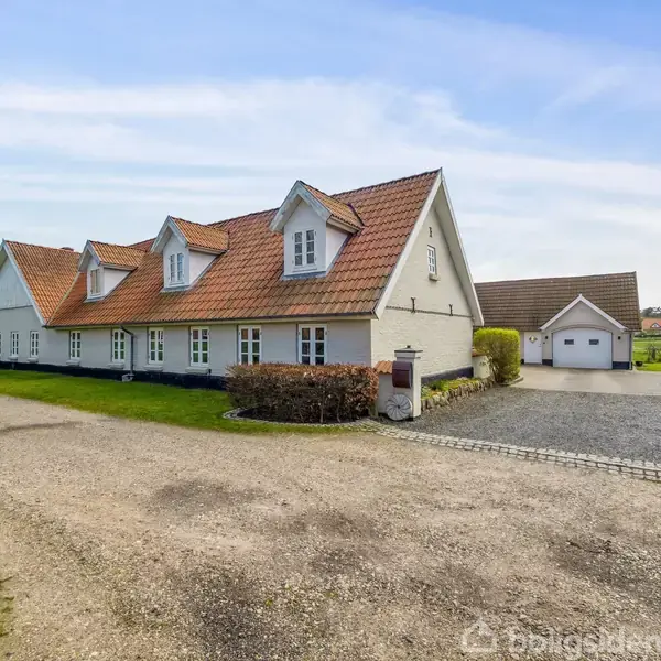 Hvide hus med rødt tegltag står ved en grusvej; omgivet af grønne marker og lidt bevoksning. I baggrunden ses en garagebygning. Himmel er klar med få skyer.