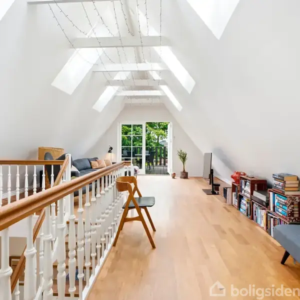 Loftstue med træbord og sofa, sofabord og reol fyldt med bøger. Skråt loft med ovenlysvinduer. Rummet er lyst med åbne døre mod en terrasse med grøn udsigt.