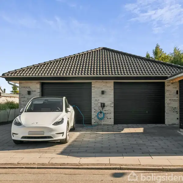 Bil oplader ved brun murstensgarage med to porte. Parkeringspladsen er flisebelagt, og omgivelserne består af andre huse og træer. Himlen er blå og skyfri.