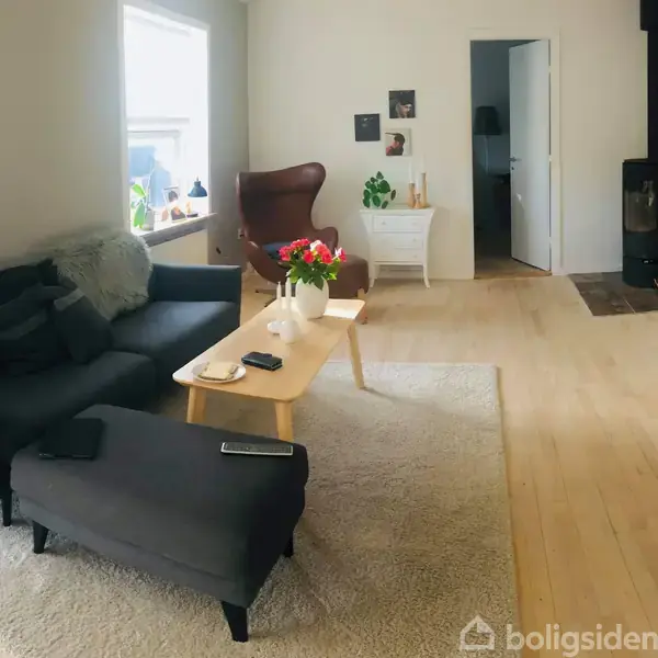 En grå sofa står i en stue ved et træbord med blomster. En lænestol er ved vinduet. Pejsen er i hjørnet. Rummet har lyse trægulve og vægkunst.