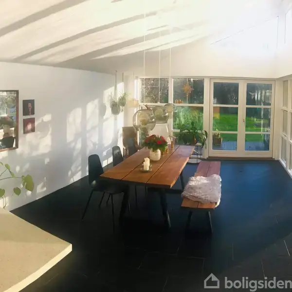 Et langt træbord står i et lyst rum med store vinduer på to vægge. Der hænger planter og billeder på væggen, og en have ses udenfor vinduerne.