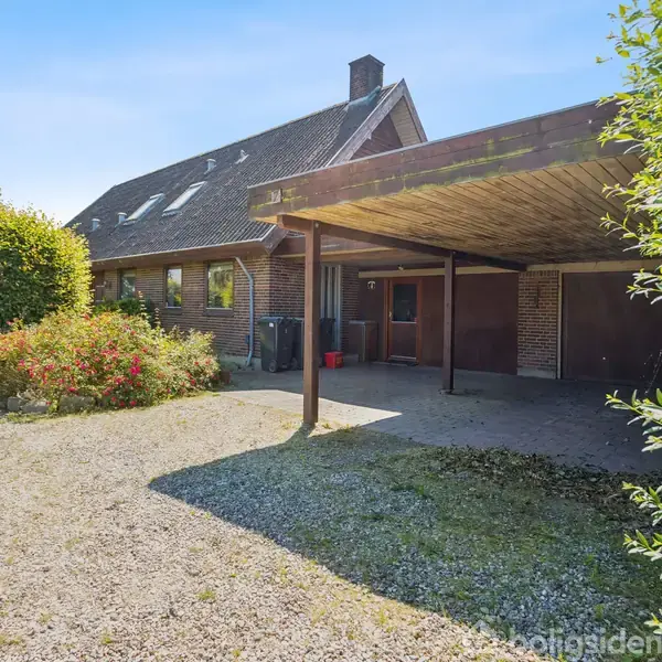 Hus med skråtag og kviste, står i en have med buske og grusbelagt indkørsel. Carporten til højre giver skygge. Omkringliggende træer giver en landlig atmosfære.