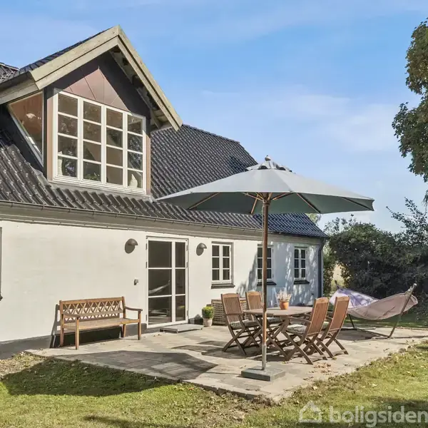 Et hvidt hus med sort tag har en terrasse, hvor der står et bord med stole og en parasol. Terrasse ligger i en have med græs og træer i baggrunden.