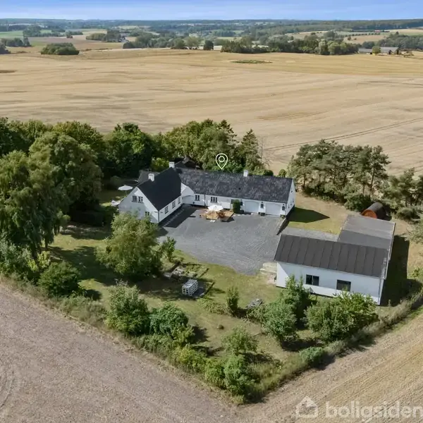 En gård omgivet af træer og marker, hvor bygningerne danner en gårdsplads. Gården er placeret i et landligt område med store åbne marker i baggrunden.