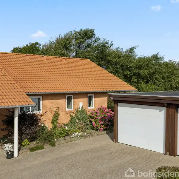 En murstenshus med rødt tegltag står ved siden af en garagebygning med to hvide porte. Omkring huset er der grønt buskads og farverige blomster. Himlen er klar og blå.
