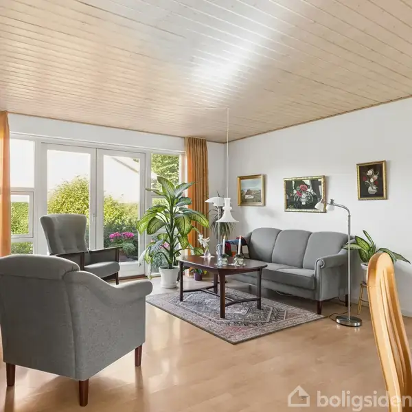 Stue med grå sofa og lænestole står omkring et sofabord på tæppe. Vinduer med orange gardiner lader lys ind. Væggene har malerier; grønne planter tilføjer liv.