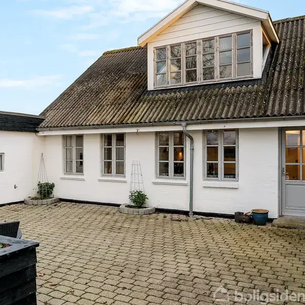 Hvid bygning med flere vinduer, står stille i en gårdhave med fliser. To døre fører ind i huset, og krukker står langs væggen. Himlen er blå.