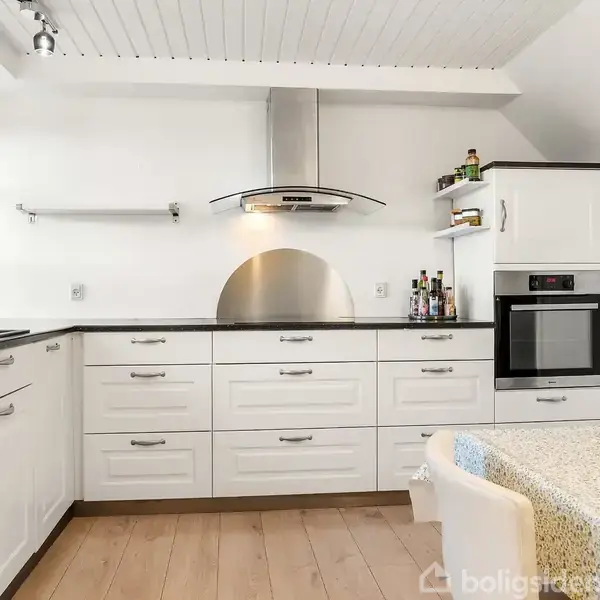 Et hvidt køkken med skabe og skuffer under en sort bordplade. En ovn og kogeplade er indbygget. Der står krydderier på hylden. Et spisebord med stole ses forrest.