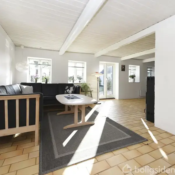 Sort sofa står i stue, omgivet af hvide vægge og store vinduer, der lader sollys strømme ind. En lys trælaminatgulv og sofabord præger rummet.
