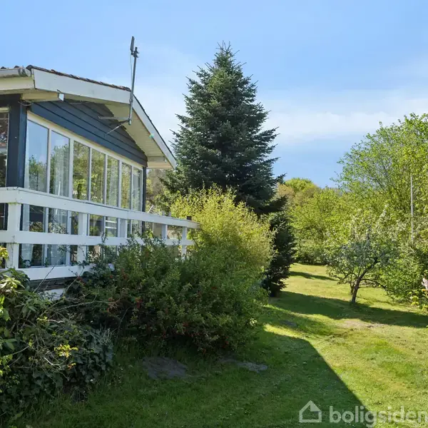 Hus med stor glasveranda omgivet af tæt have med grønne buske og træer på en solrig dag. Mærkbar fredfyldt atmosfære med blå himmel i baggrunden.