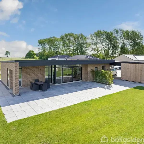 En moderne bungalow står stille på en græsplæne med solrigt vejr. Terrassen har et havebord og stole, omgivet af en velholdt have og skov i baggrunden.