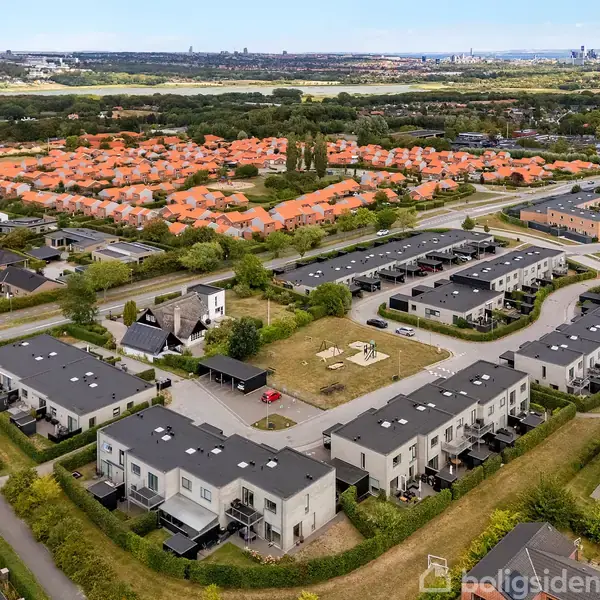 Rækkehuse med flade tage ligger langs en snoet vej. I baggrunden ses en klynge af enfamiliehuse med røde tage. Området er grønt med træer og marker omkring.