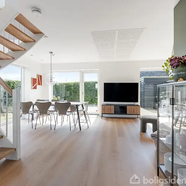 Trætrappe fører op i moderne stue med spisebord og stole. TV står ved vindue, omgivet af have. Skænk med glaslåger viser service. Lyserum med minimalistisk indretning.