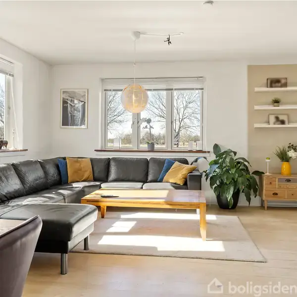 Stor, sort lædersofa står i en lys stue fyldt med naturligt lys fra flere vinduer. En træbord, gulvtæppe og planter skaber en hyggelig atmosfære.