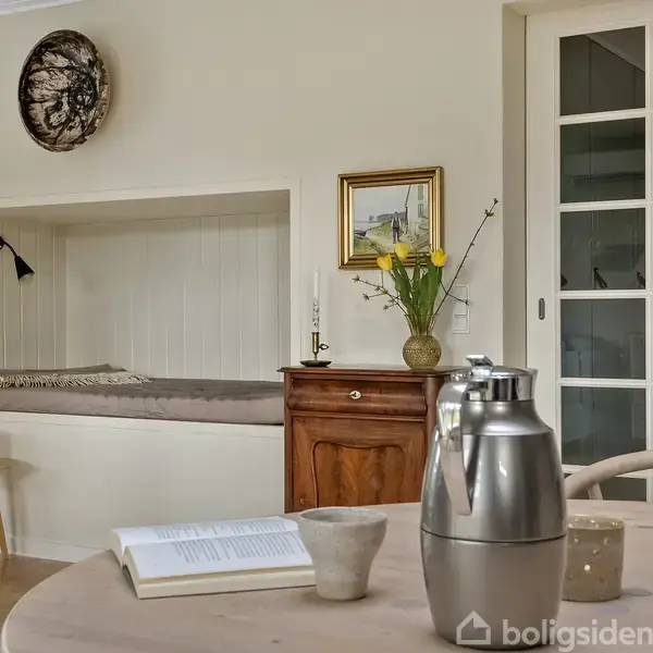 En stue indeholder et rundt bord med kaffekande, kopper og en åben bog. I baggrunden ses en indbygget hyggekrog, en trækommode med vase og maleri samt en glasdør.