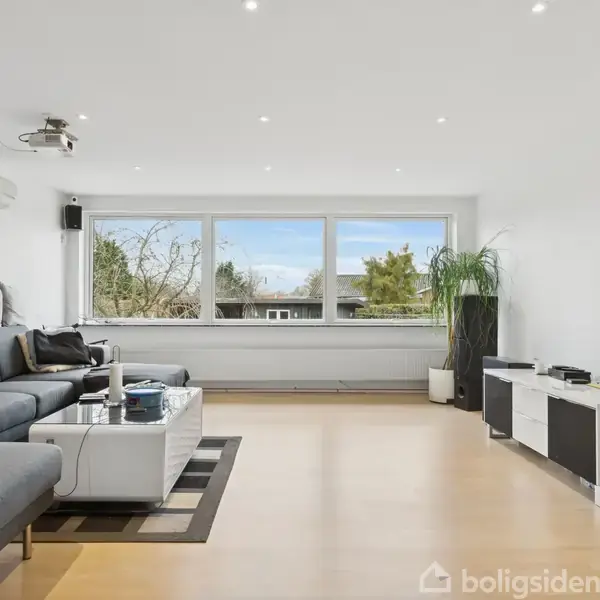 En stor stue med en grå hjørnesofa står til venstre. Et sofabord og underholdningsreol ligger på højre side, med projektor i loftet og stort vindue i baggrunden.