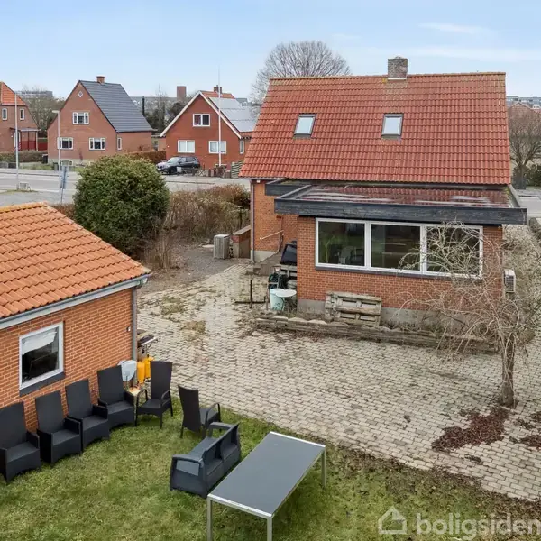 To murstenshuse med røde tage og en gårdhave. I haven står sorte havemøbler og et bord på en græsplæne. Omkring er flere lignende huse.