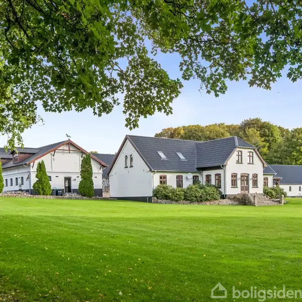 Et hvidt, landligt hus med sort tag står i en frodig have med græsplæne. Bagved ses flere bygninger og træer i en skovklædt baggrund under en blå himmel.