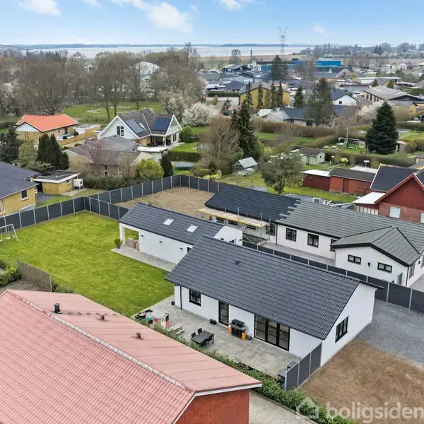 Hvide huse med sorte tage i en rolig forstad; set fra luften. Store grønne haver omgiver husene, mens kvarteret viser en række spredte træer og flere huse i baggrunden.