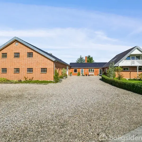 To murstensbygninger står stille omkring en grusbelagt gårdsplads, med grønne buske og træer omkransende. En bygning har en altan med tag, mens den anden har mindre vinduer.