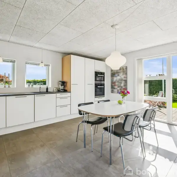 Et moderne køkken med hvide skabe og apparater står åbent mod en terrasse. Et rundt bord med sorte stole er i centrum. Store vinduer viser en grøn have.