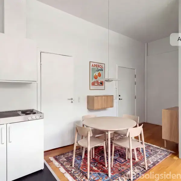 En lille spisekrog med et rundt træbord og fire stole står på et farverigt tæppe. Baggrunden indeholder et minimalistisk køkken og en plakat med teksten "Aperol Spritz".