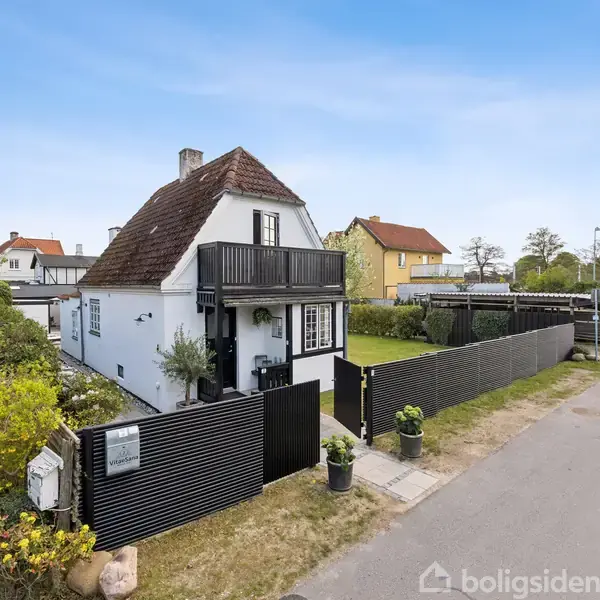 Hvidt hus med rødt tag står ved vej, omgivet af sort stakit og have. Der er en balkon på første sal. Biler parkeret i baggrunden og andre huse i nærheden.