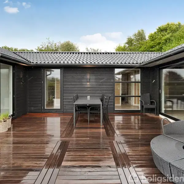 Sortmalet træterrasse med blanke træplanker og moderne, udendørs møbler. To glasdøre åbner ind til huset. Grønne træer udgør baggrunden under en blå himmel med enkelte skyer.