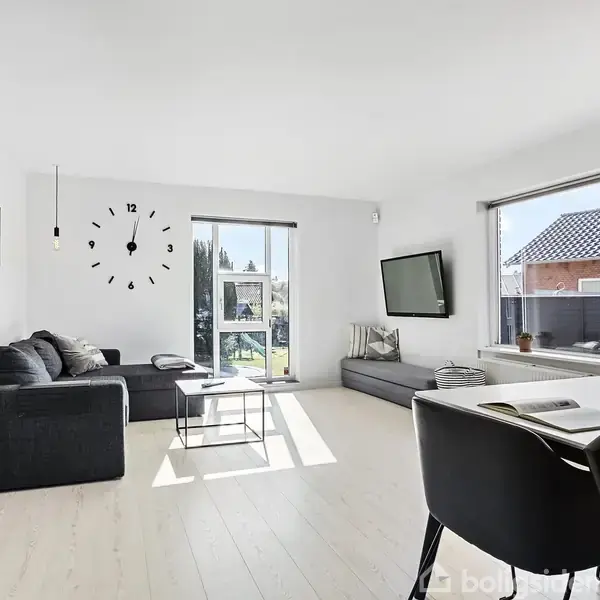 Sort sofa står i en lys stue. En vægmonteret TV hænger ved siden af et stort vindue, der viser en have. Et spisebord med stole ses i forgrunden. Vægur uden visere.