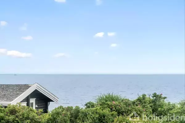 Et hus med gråt tag står stille ved kanten af vandet omgivet af grønne buske, mens havet strækker sig mod horisonten under en klar, blå himmel.