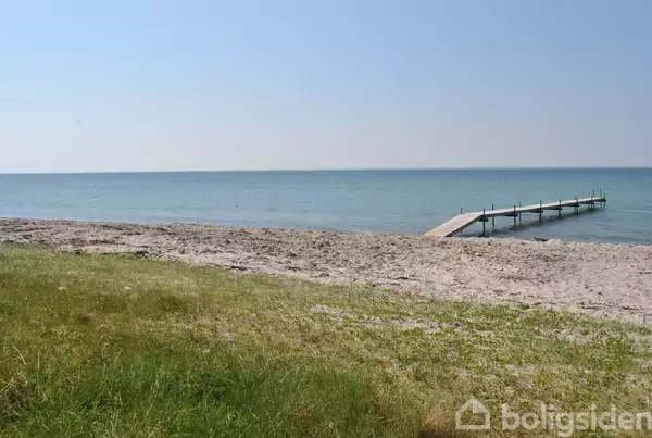 En træbro strækker sig ud over det blå hav. Stille strand med sand og græs omgiver området under en klar blå himmel.