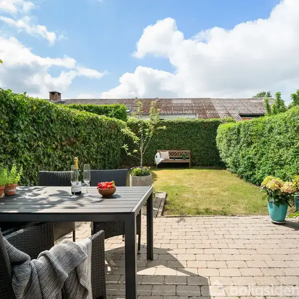 Et havebord med stole står på en flisebelagt terrasse. Der er en flaske vin og skål med jordbær på bordet. Haven er omgivet af grønne buske under en blå himmel.
