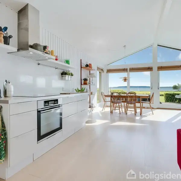 Køkken med moderne hvide skabe og integreret ovn, beliggende i et lyst rum med spisebord og stole i baggrunden. Store vinduer giver udsigt til en terrasse og have.