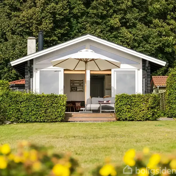 Et hvidt sommerhus med åben dør står omgivet af grønne buske og græsplæne, mens en parasol skygger terrassen. Skoven danner baggrund.
