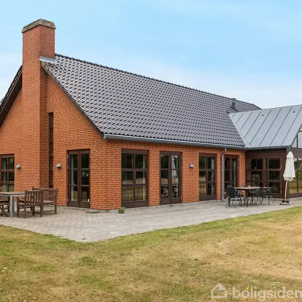 Huset er en rød murstensvilla med sort tegltag, placeret i en rummelig have. Der står havemøbler på en terrasse af fliser ved husets side.