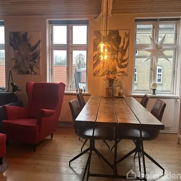 Rummelig stue med en stor træbord omgivet af stole, en rød lænestol ved siden af et vindue og reoler fyldt med bøger. Stjerner hænger i vinduerne.