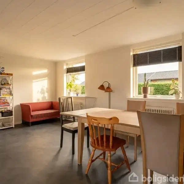 En rummelig stue med spisestue har et langt bord omgivet af forskellige stole. I hjørnet står en rød sofa nær en bogreol. Vinduer bringer lys ind i rummet.