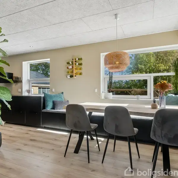 En moderne stue med store vinduer viser en spiseplads med fire grå stole omkring et bord. En grøn plante står i hjørnet, og dekorationer hænger på væggen.