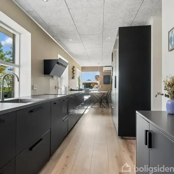 Moderne køkken med sorte skabe, en indbygget ovn og en stor vask. Vindue med udsigt til grønne træer. Trægulv og lyst loft, blomstervase på skab.