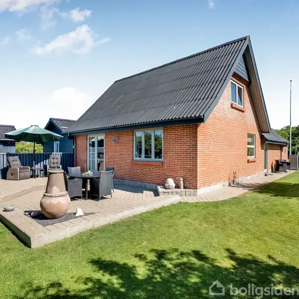 Rødt murstenshus med sort tag står på en velplejet græsplæne. En flisebelagt terrasse har havemøbler og en terracotta-ovn. Solrig dag og træer i baggrunden.
