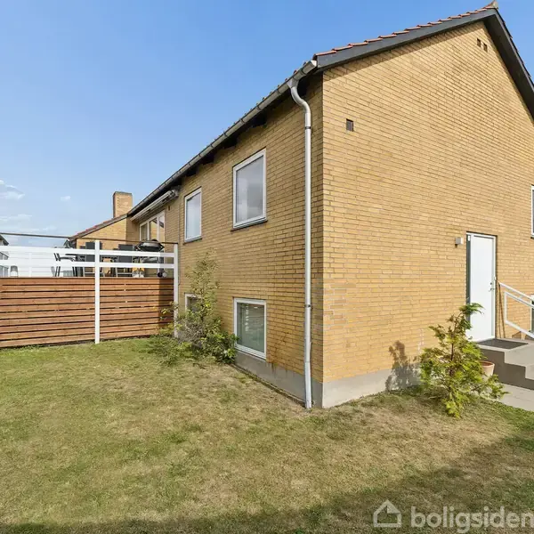 Et gult murstenshus med en hvid dør og flere vinduer. Husets side har en tilhørende hævet terrasse med gelænder. Det står i en grøn have under en blå himmel.