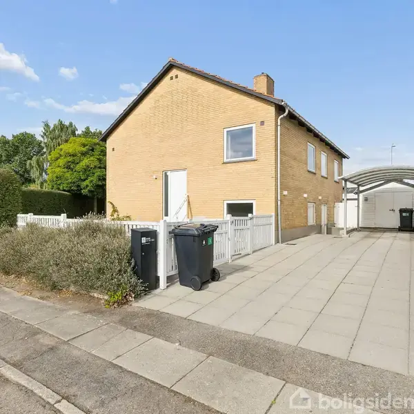 Et toetagers gult murstenshus står ved en flisebelagt indkørsel med affaldscontainere. Omgivet af hække og træer i et boligområde, med carport i baggrunden.