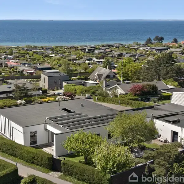 Et moderne, hvidt hus med fladt tag ligger i en velholdt have. I baggrunden ses en kystlinje og hav med små bølger. Omkringliggende huse udgør et villakvarter.