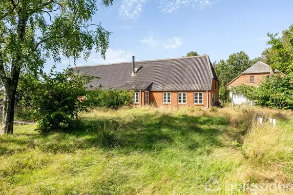 Rødt murstenshus med skrå tag står i solrigt græsareal omgivet af buske og træer, hvilket skaber en fredfyldt, landlig atmosfære.