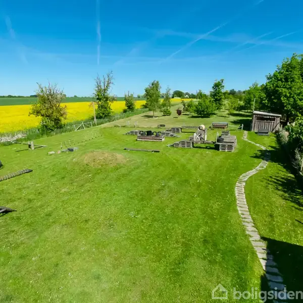 Et gårdhaveområde med to bygninger omkring, hvor den ene bygning har et udendørs siddeområde. Der er også en vandpumpe og diverse havemøbler i solen.