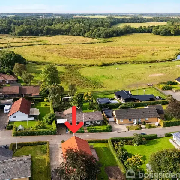 Rødt hus med markeret rød pil over taget, omgivet af andre huse og grønne områder. Baghaven grænser op til åbne enge med en lille bæk i baggrunden.